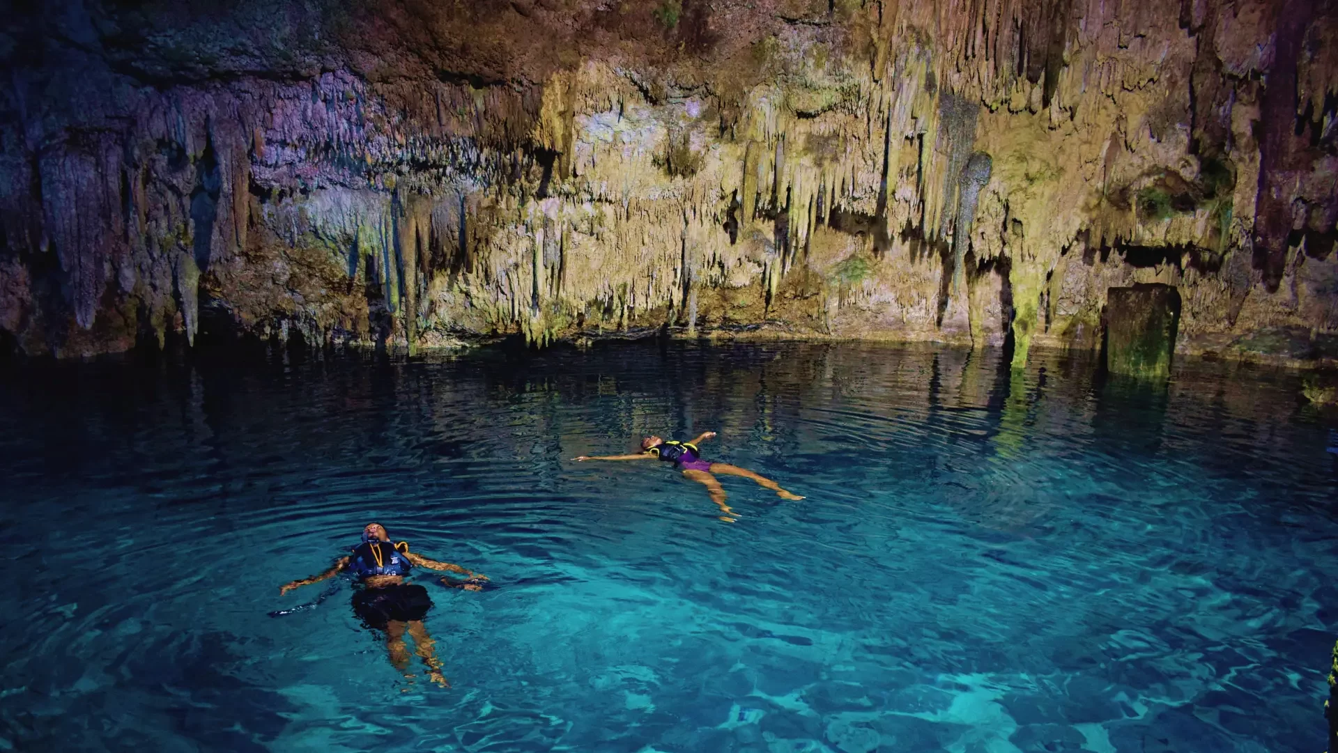 Two swimmers relax in the clear waters of a cenote, surrounded by limestone formations, during a private tour by Carey Tours.